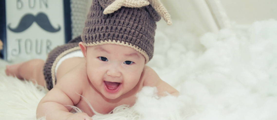 a happy chinese baby with a cute beanie on smiles atop a sheepskin rug, representing our discussion regarding chinese babies and eczema in china